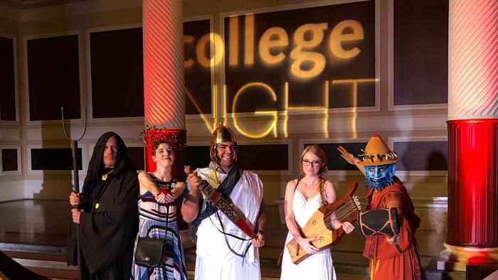 Five people in medieval costumes stand before a light projection reading College Night
