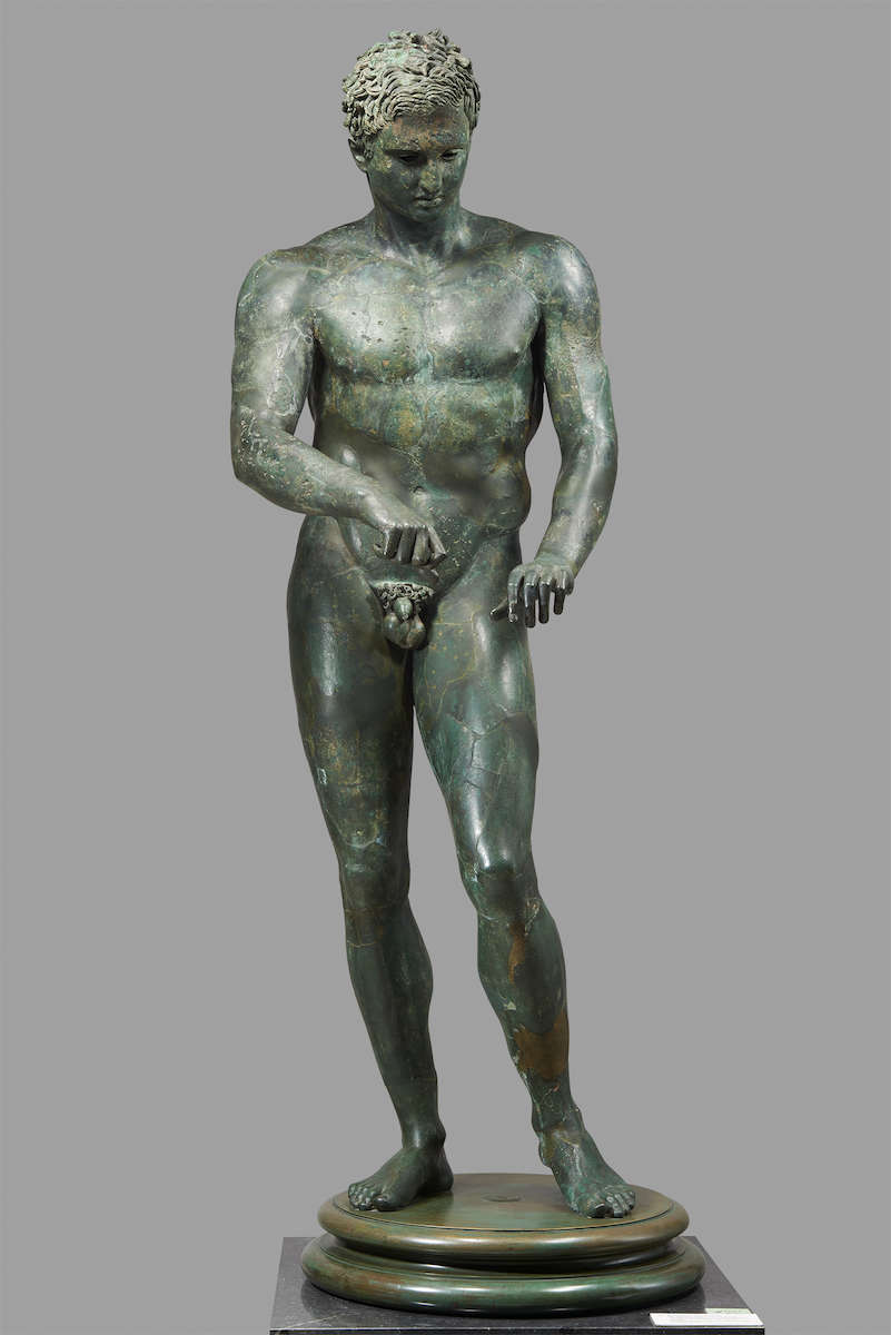 Bronze sculpture of standing nude male, short hair, looking down with his arms raised slightly in front of him and his hands poised as if in action.