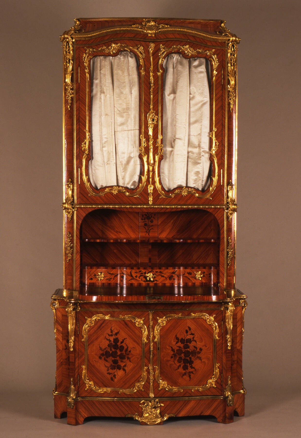 a tall secretary desk with two sets of doors, one under the desktop of shelves and drawers, and the other above, constructed with three different types of wood and decorated with gilt bronze mounts, white marble accents, and two white satin curtains installed inside the upper set of cabinet doors