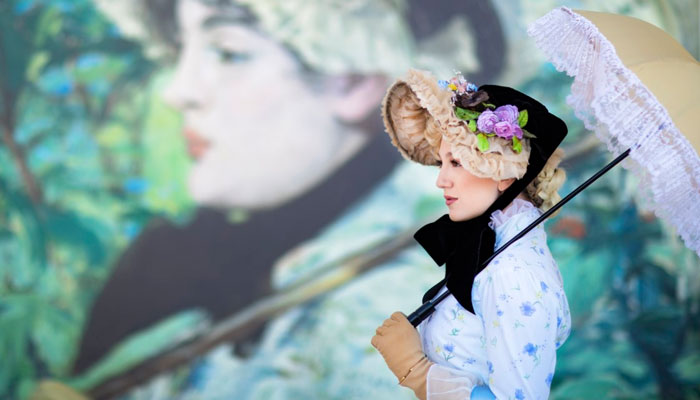  A woman dressed in clothing from the 1800s holds a parasol in front of the painting 