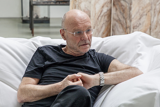 Middle-aged white man with glasses (artist Anselm Kiefer) sits on a large cushion, interlocking his fingers as he looks off-frame to his left.