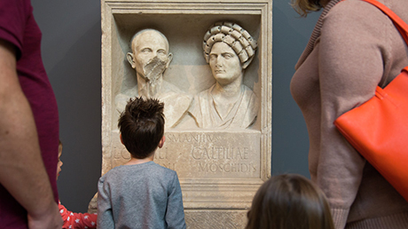 A child and his family look at two stone busts of a woman and a man with half a face.