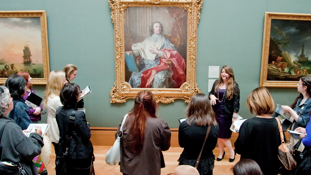 An older man leans down pointing at part of a sculpture to several young children looking intently