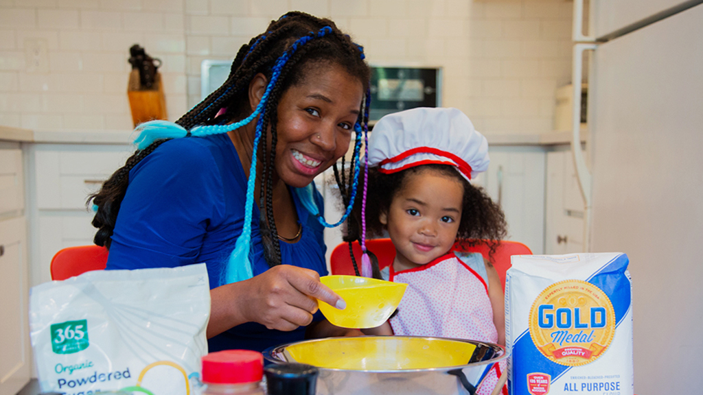A woman and a young girl are in a kitchen smiling at the camera while mixing together powdered sugar and flour in a bowl