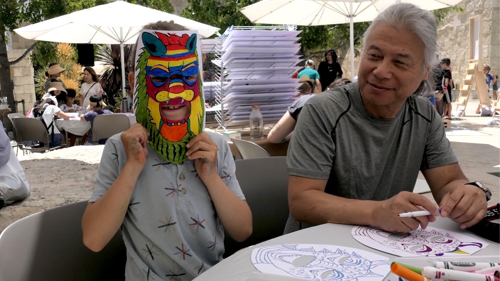 A child sitting at a table holds a colored mask in front of his face while an adult smiles at him