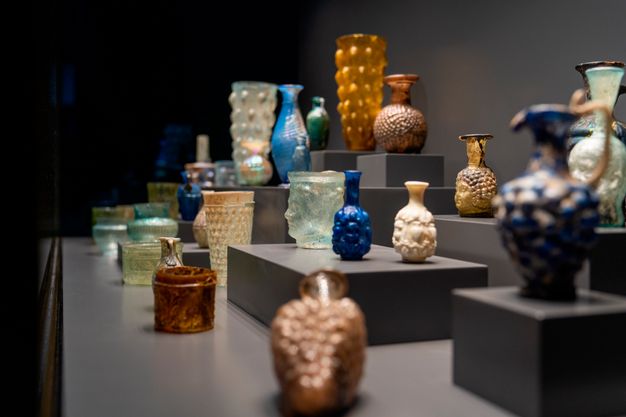 A museum gallery with dark walls and ceiling-mounted spotlights contains multiple display cases filled with ancient glass vessels. The central case holds a single artifact, while the surrounding cases feature grouped arrangements.