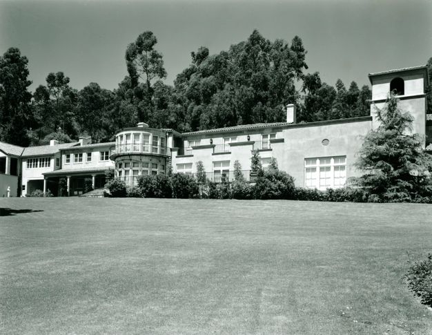 Getty Ranch House features a large hillside residence with a central rotunda-style window structure, an arched entrance, and a tiled roof. Surrounded by a manicured lawn and tall trees, the house blends modern and Mediterranean elements.