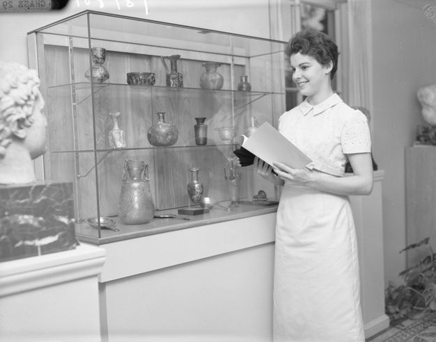 A woman in a white dress holds an open book while standing beside a glass display case containing ancient vessels. A marble bust mounted on a pedestal appears in the foreground.