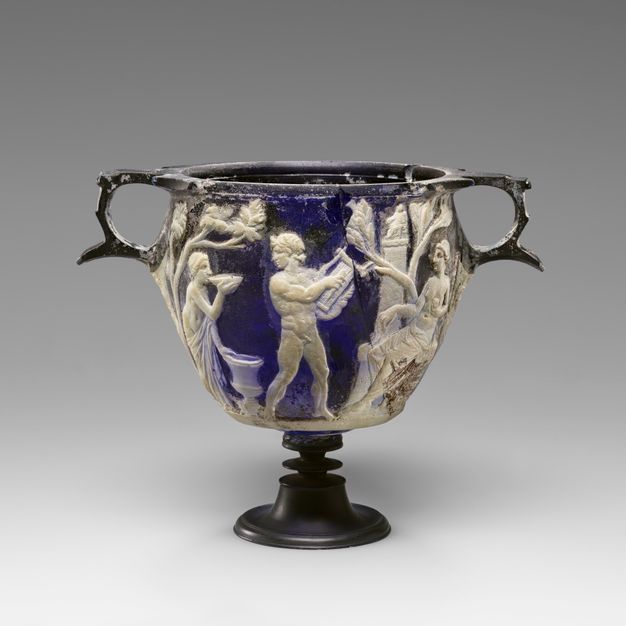 A two-handled blue vessel with a flared rim and footed base features opaque white relief figures. A male figure with a lyre stands at the center, flanked by draped female figures.