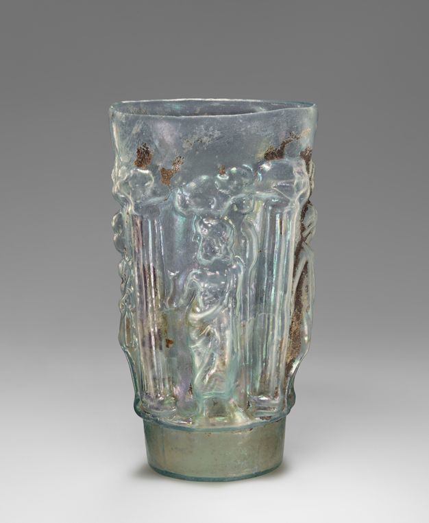 A translucent beaker with an iridescent surface shows standing figures framed by architectural columns. Weathering and encrustation appear across the surface.
