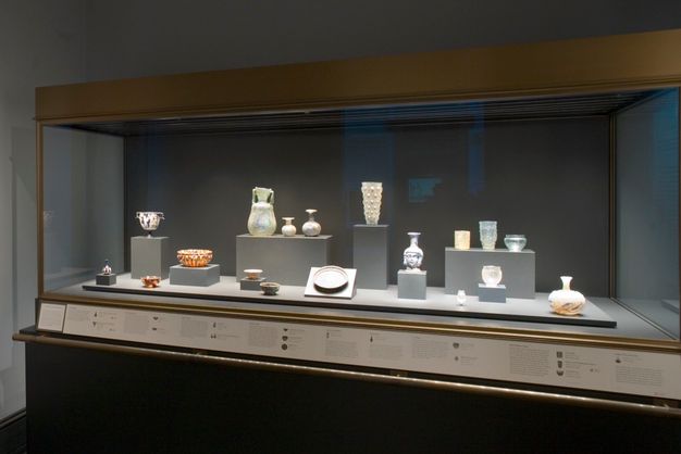 A museum display case contains ancient glass vessels of varying shapes, sizes, and hues. Objects include pitchers, bowls, goblets, and flasks, arranged on tiered platforms with labels.