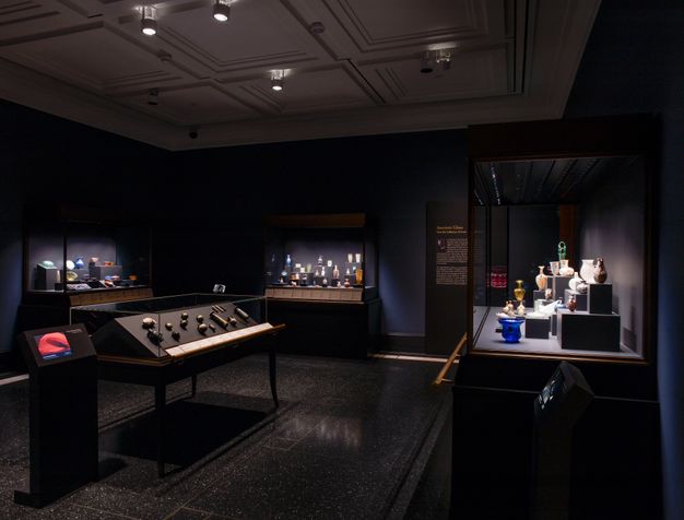 A dimly lit museum gallery features multiple display cases containing ancient glass vessels and artifacts. Spotlights illuminate the exhibits, which are arranged on raised platforms with accompanying text panels.
