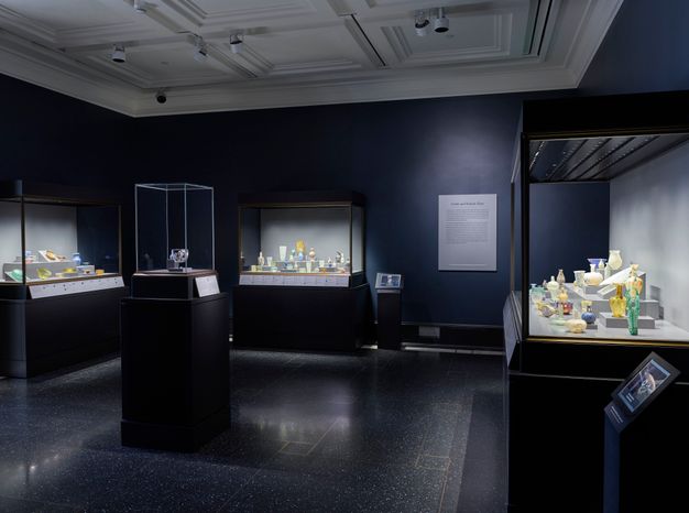 A group of ancient glass vessels is arranged on tiered black platforms inside a display case. The vessels vary in form, texture, and hue, including ribbed, globular, and flared shapes.