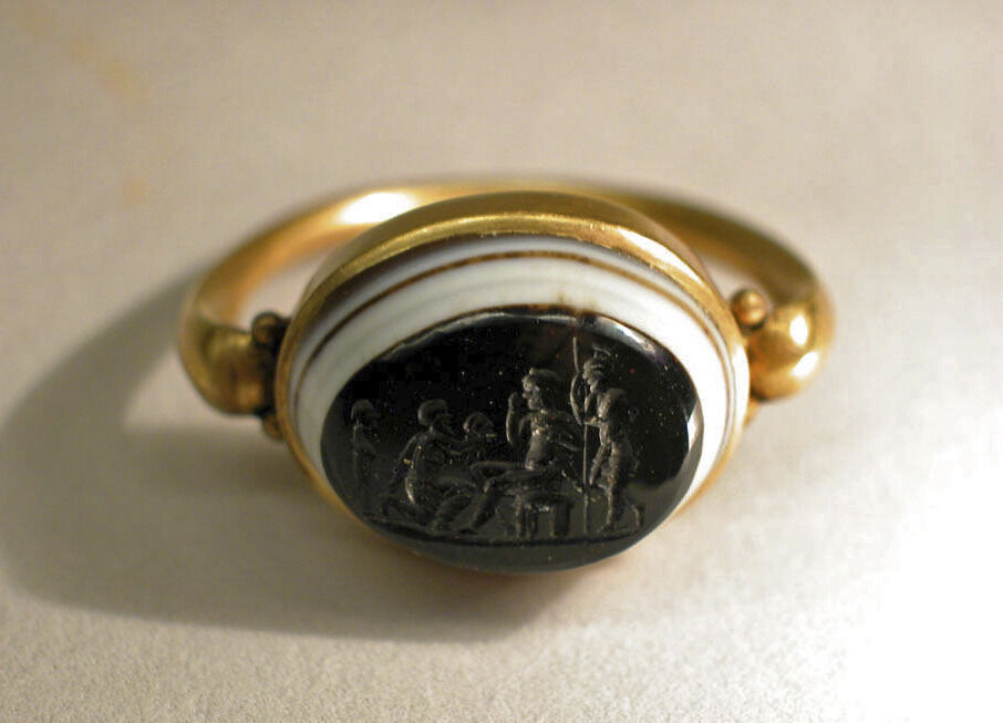 Black, oval gem inset in a golden ring.  The gem depicts a recessed scene in which a kneeling individual offers a severed head to a person who sits before them. Two individuals appear standing, each behind one of the central figures.