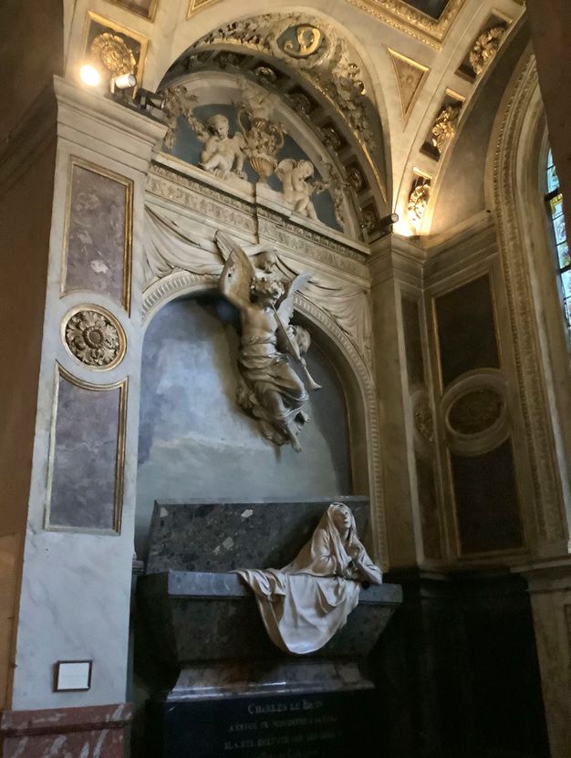 Photograph of a tomb placed inside a church. It features a sarcophagus in black marble and with the lid open from which a draped human figure carved in white marble seems to be coming out. A seminude angel with a trumpet hangs above. The composition is topped with a human skull.