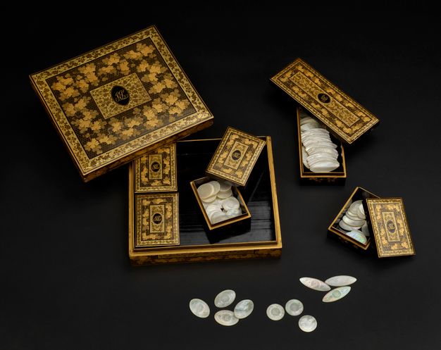 Lacquer square box with the lid open. Inside there are three smaller boxes that match the bigger one in their ornamentation: two closed and one open. Two additional matching boxes are placed outside the bigger one with their lids also open. The open boxes show their contents. These are mother-of-pearl counters of different shapes.