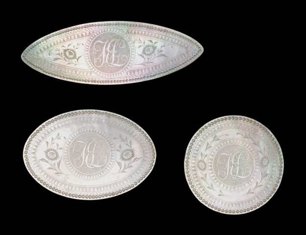 Three mother-of-pearl counters against a black background. The one on the top row is shuttle-shaped. On the bottom row, the one on the left is oval and the one on the right is rounded. The three counters feature in the center the initials J, E, and L surrounded by floral motifs.
