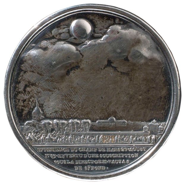 Rounded silver medal depicting the ascent of a hot-air ballon. The ballon is shown at the top, floating among clouds. Below, there is a crowd of people standing in front of buildings and trees. Four lines of text in French appear at the bottom of the medal.