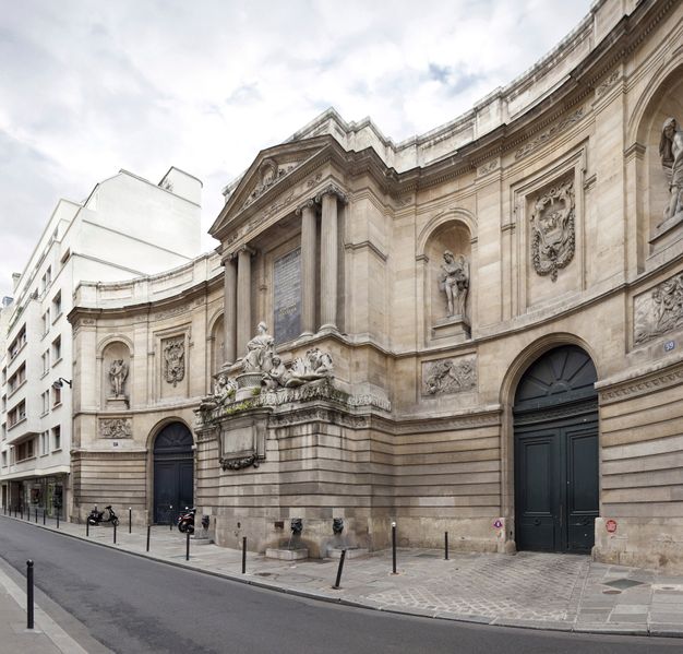 White structure resembling the façade of a palace in a neoclassical style. The central section protrudes and includes a group of three freestanding sculptures of human figures in front of a frontispiece with a triangular pediment. This section is flanked by two curved wings with statues in niches and bas-relief panels. It features spouts in the shape of an animal’s head at the street level.