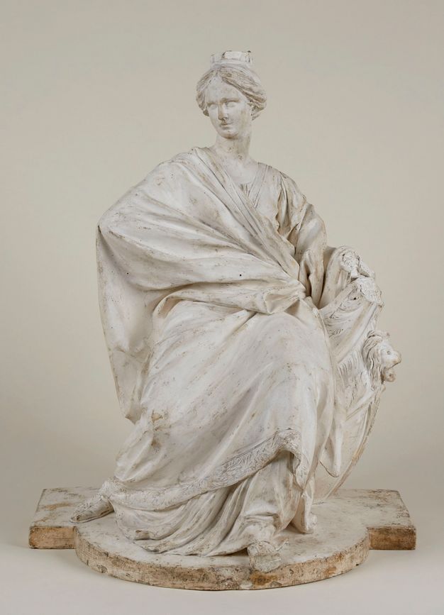 Plaster sculpture depicting a seated woman. Her entire body is covered in drapery that hides her shape, only leaving uncovered her head and her feet. Her hair is tied in the back and adorned with a crown.