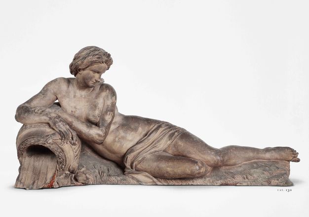 Sculpture of a nude woman reclining over an urn from which water emerges. The sculpture looks cream to grey in the surface, but a fracture reveals a clay core.