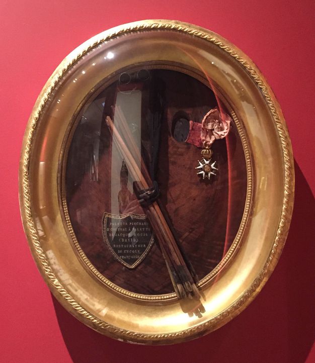 Framed and encased collection of objects, including a palette, brushes, and a military decoration. A piece of leather in the shape of a shield with an inscription in gold is attached to the surface of the palette.