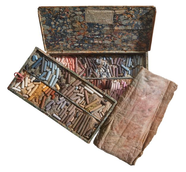 Open box featuring two trays filled with pastel sticks roughly arranged by color. The lid is lined with marbled paper. A padded rectangle of fabric stained with pastel powder appears to the right of the box.
