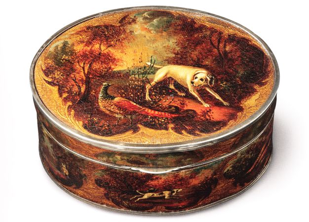 Circular metal box decorated with papier maché around its body and on the top of the lid. The scene on the lid shows a dog and bird in a forestry environment. A dog can also be seen on the sides of the box with its legs outstretched.
