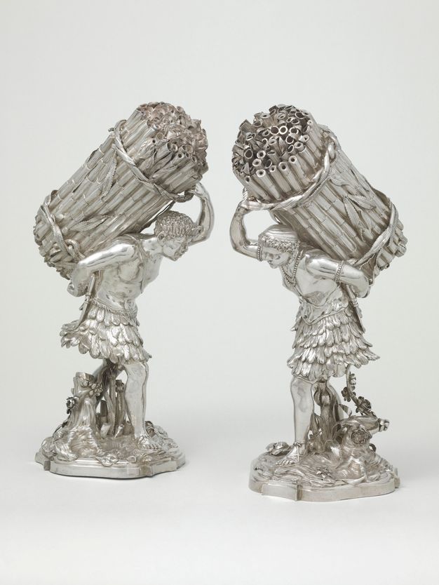 Two silver casters in the shape of men carrying large loads of sugar cane. They both lean forward to accommodate the bundles on their back, while one of their arms reaches back and towards the bottom of the load, and the other reaches up, above their heads, to hold the upper portion of the load. The top of some of the sugar cane stalks are perforated. Both individuals are shown wearing feathered skirts and sandals. The person on the caster to the left also wears a necklace and armlets.