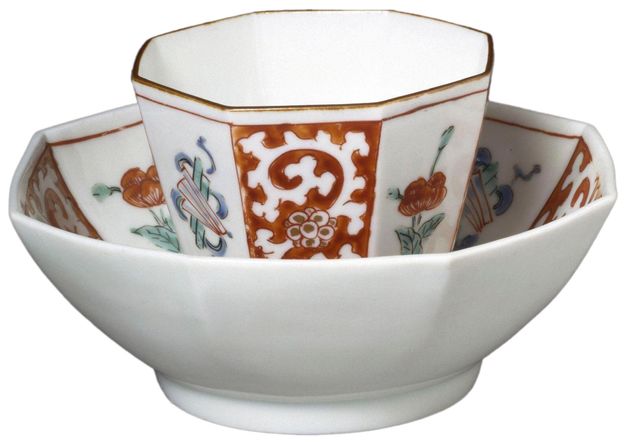 Porcelain octagonal cup placed inside an octagonal saucer. The saucer is bare of any decoration on its outside but features floral motifs in its interior in orange, blue, and green. No handle can be seen on the cup from the angle in which it was photographed. The cup features a golden rim and floral decoration on its outside in the style and colors as the saucer. The interior of the cup appears to be plain white.