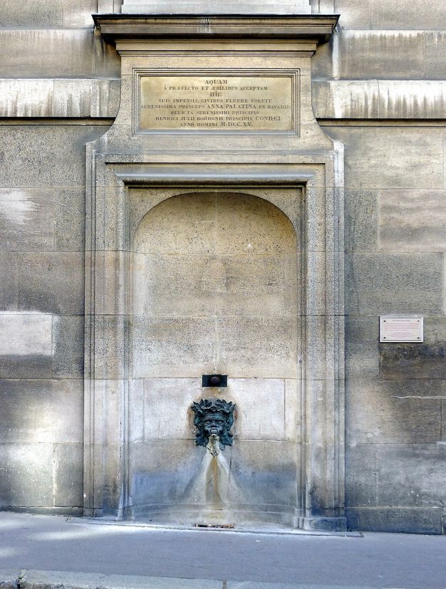 Fountain featuring a spout set in a bronze mascaron and placed in a recessed stone niche. A plaque with an inscription in Latin hangs above.