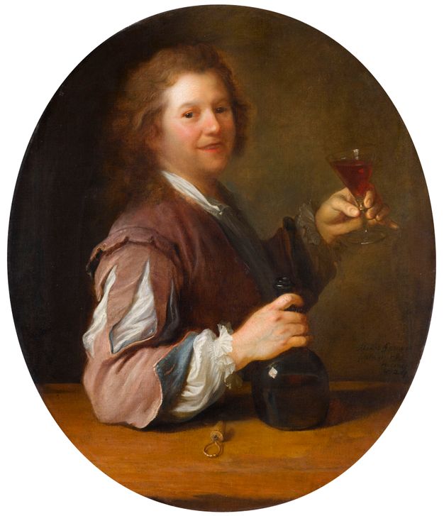 Portrait of a man from across a wooden table. He is shown raising a wine glass with his left hand while grasping the neck of a green wine bottle that rests on the table. A cork can be seen next to the bottle.