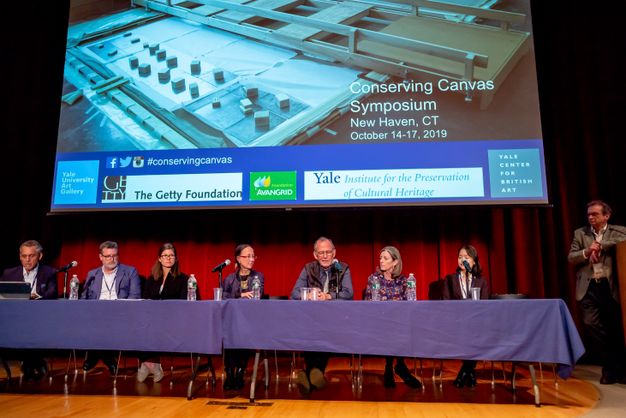 A panel of speakers sits behind a table below the presentation screen, which shows the symposium title.