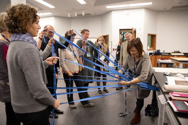 People stand holding blue ribbons attached to a table. A woman stands near the table to weave the ribbons together.