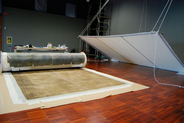 Photograph of painting lining lain on a wooden floor within a conservation room. A metal lining stretcher is suspended to the wall with rope and hangs above the lining.
