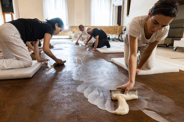 Medium photograph of conservators kneeling atop the back of a painting and applying adhesive.