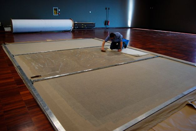 Wide photograph of a conservator kneeling atop the back of a lining canvas and applying adhesive.