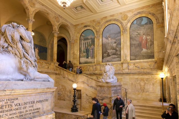 Wide photo of a marbled staircase. Lion statues frame the stairwell and the mural in figure 12.1 is on the wall behind.