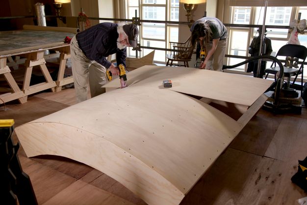 Wide photo of two conservators drilling into a curved wooden structure.