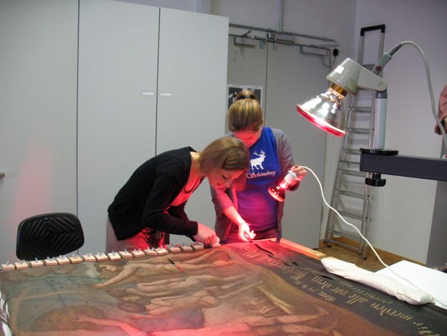Photo of two conservators further repairing the painting under red light.