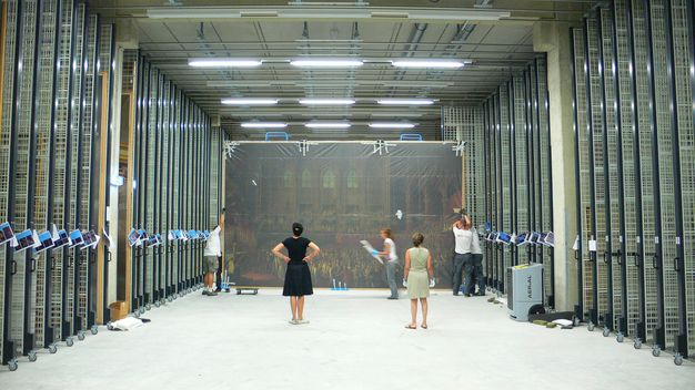 Wide photo of people storing a large painting within a museum vault.