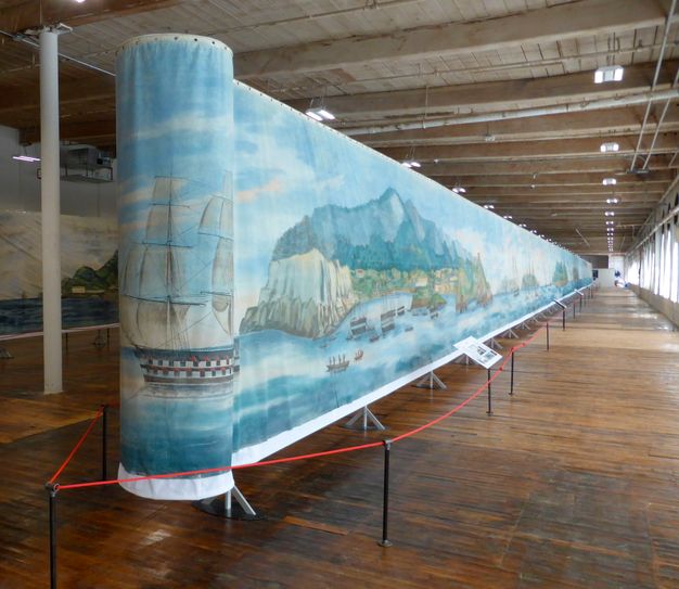 Wide photo of a long, wood-paneled exhibition room. A long seascape painting canvas stands at the center of the room.