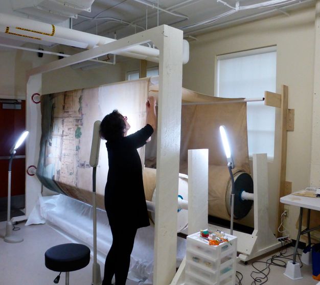 Photo of a conservator in black clothes treating the canvas (figure 23.1), which is rolled up.