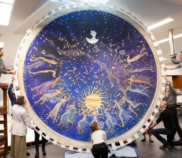 Wide photo of people in a conservation lab holding up a large, blue, circular painting.