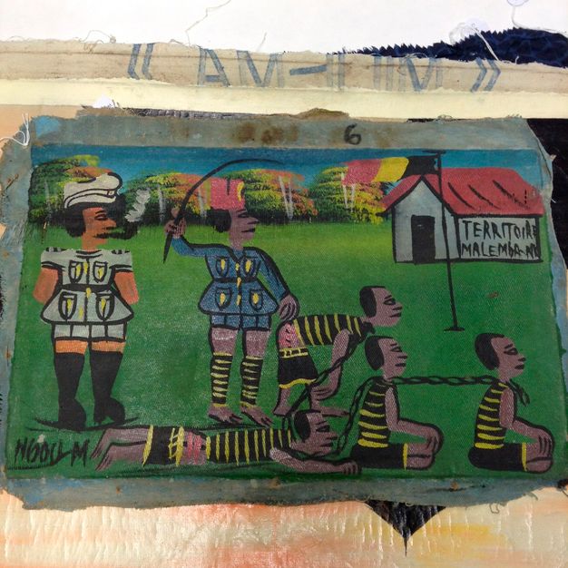 Photo of a painting canvas atop a table. The painting depicts several human figures atop grass, with four figures chained around the neck. Two figures who appear to be guards stand above them.