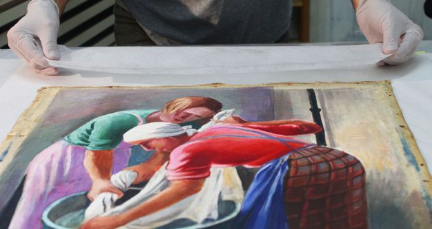 An oil painting on canvas lying flat on a white sheet. A pair of hands in latex gloves stretches a mesh strip over the sheet at the top of the painting.