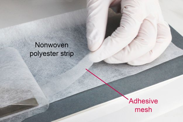 A gloved hand delicately places a strip of adhesive mesh on top of a larger, nonwoven polyester strip.