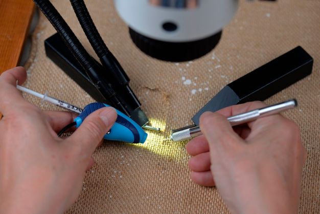 A pair of hands working on canvas, the left hand using the syringe-like applicator of the Consolidation Pen Winnie and the right hand holding a pointed tool.