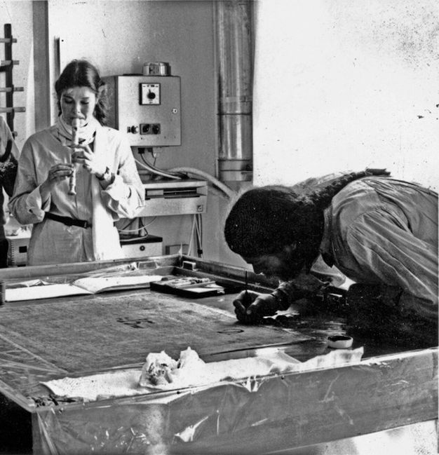 Black and white photograph of a man (right) leaning over a table and painting with a woman (left) playing the recorder.