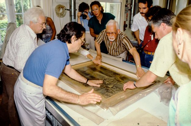 Photo of a group of people leaning over a table to examine a painting.